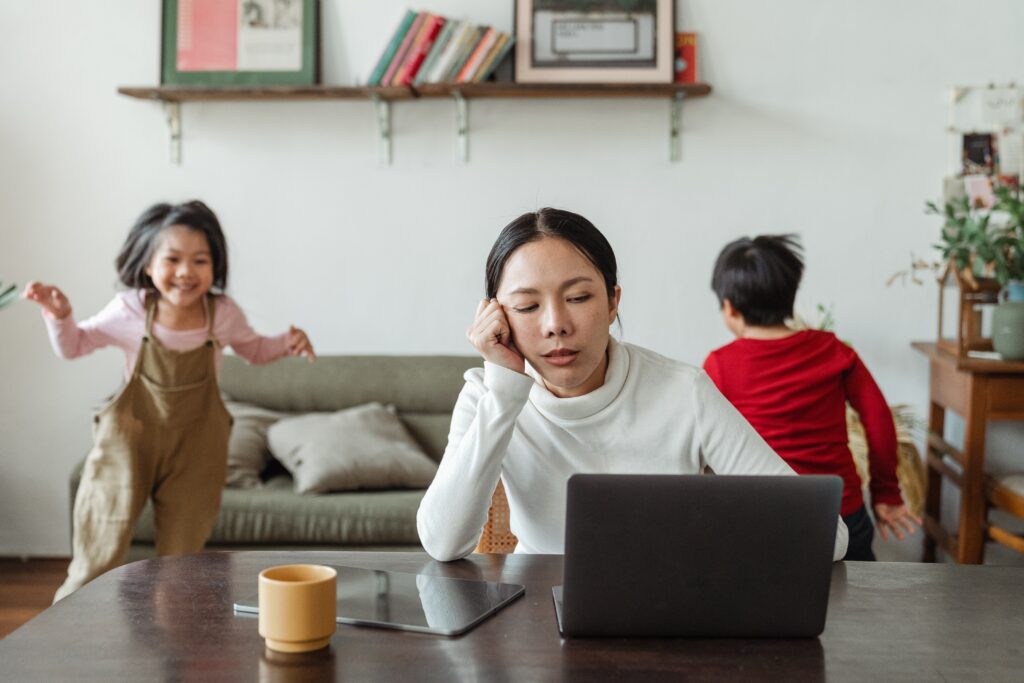 woman at desk stressed with two children running around in the background